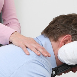 Ein Mann in Businesshemd liegt mit seinem Kopf auf einem mobilen Massagestuhl. Vom linken Bildrand aus berührt eine Hand ihn im Schulter- und Nackenbereich