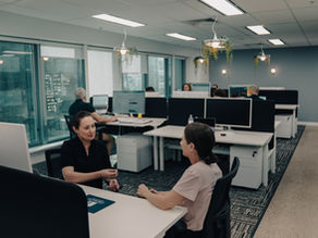 An office with people sitting at desks workign on monitors in the background and 2 women talking in the foreground of the office