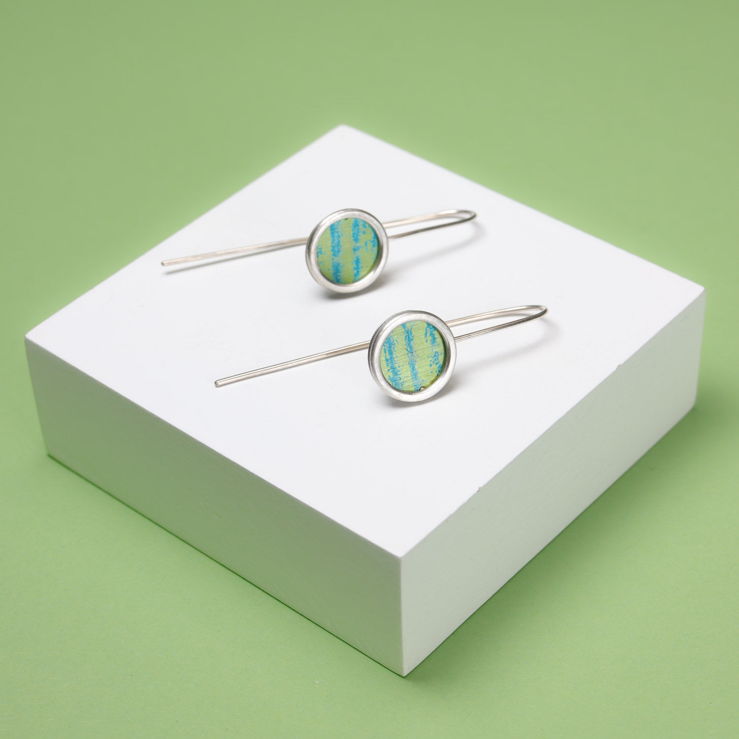 Green and turquoise stripe silver drop earrings on a white plinth on a green background