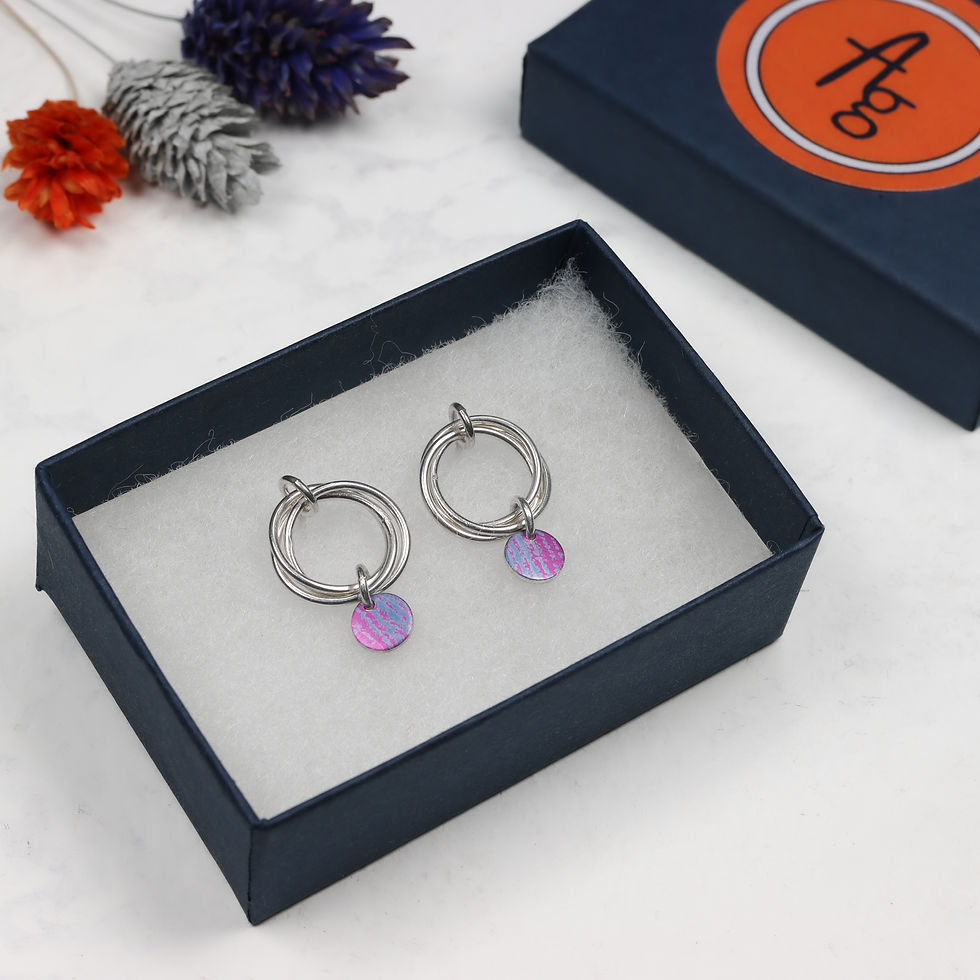 Thumbnail: Magenta pink and mauve blue aluminium and 3 silver ring earrings in an open dark blue jewellery box with lid and flowers behi