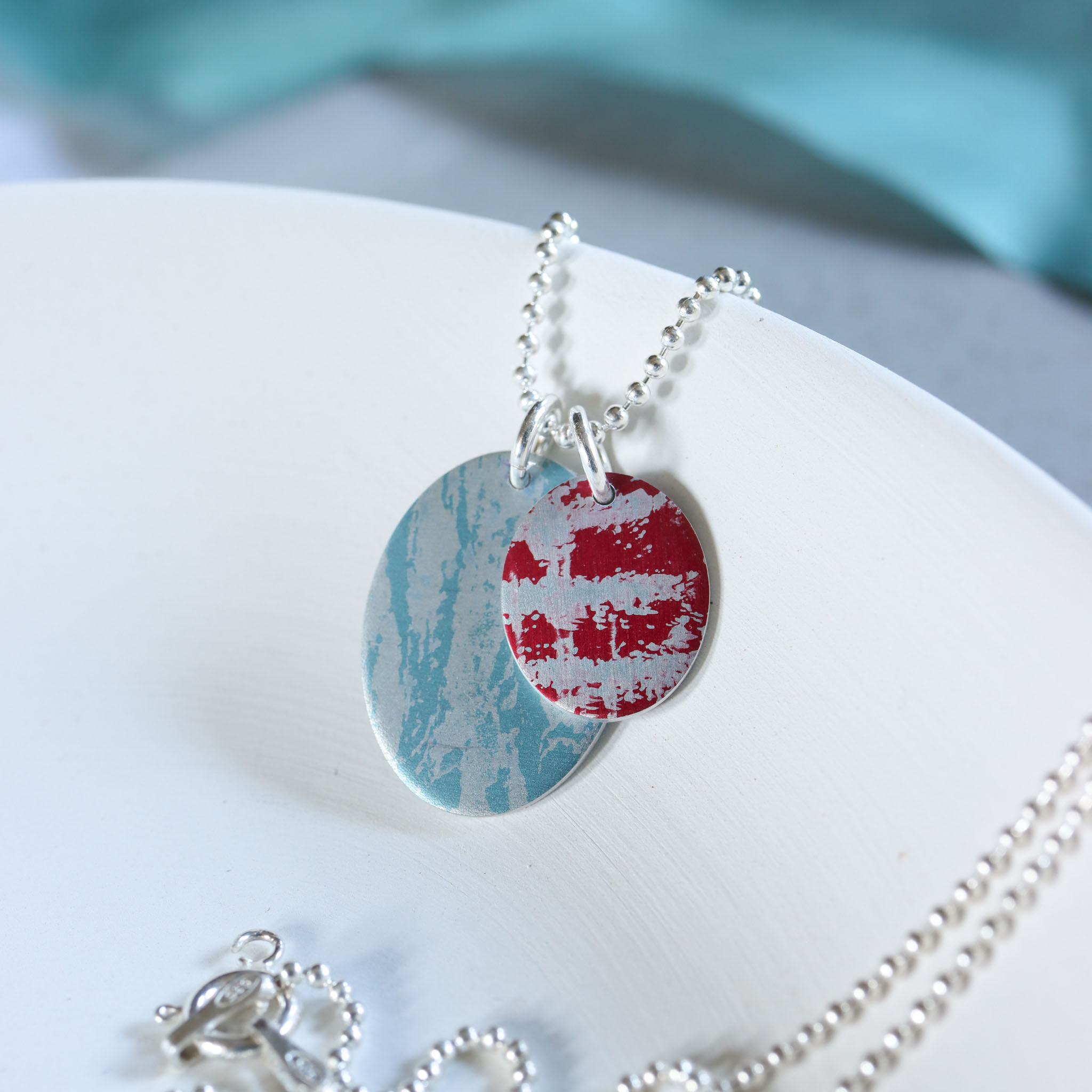 Two ovals one light blue, one red on a silver chain in a white trinket dish with turquoise blue ribbon behind