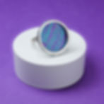 Purple and turquoise aluminium and silver rivet ring on a white round plinth on a purple background