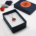 Thumbnail: Dark orange sea glass and silver pendant necklace in an open dark blue jewellery box with lid and flowers behind