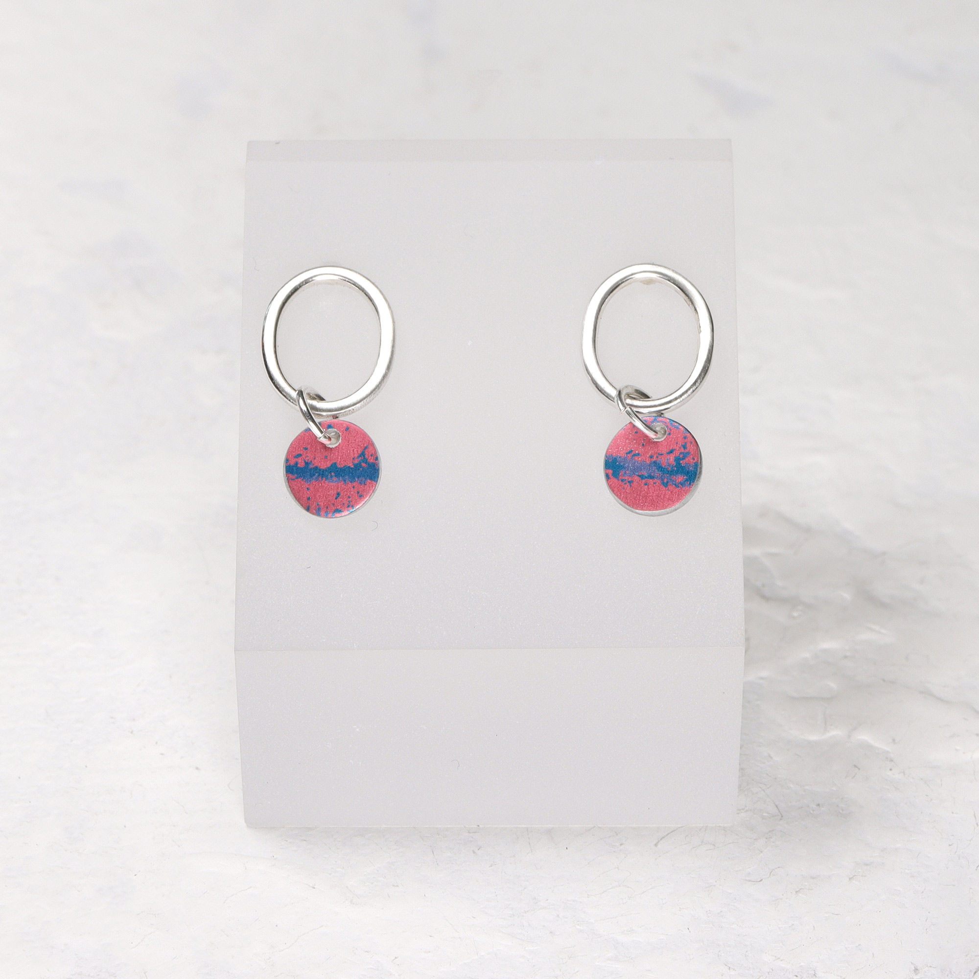 Pink and blue aluminium mini drop silver oval stud earrings on a white opaque stand on a white background