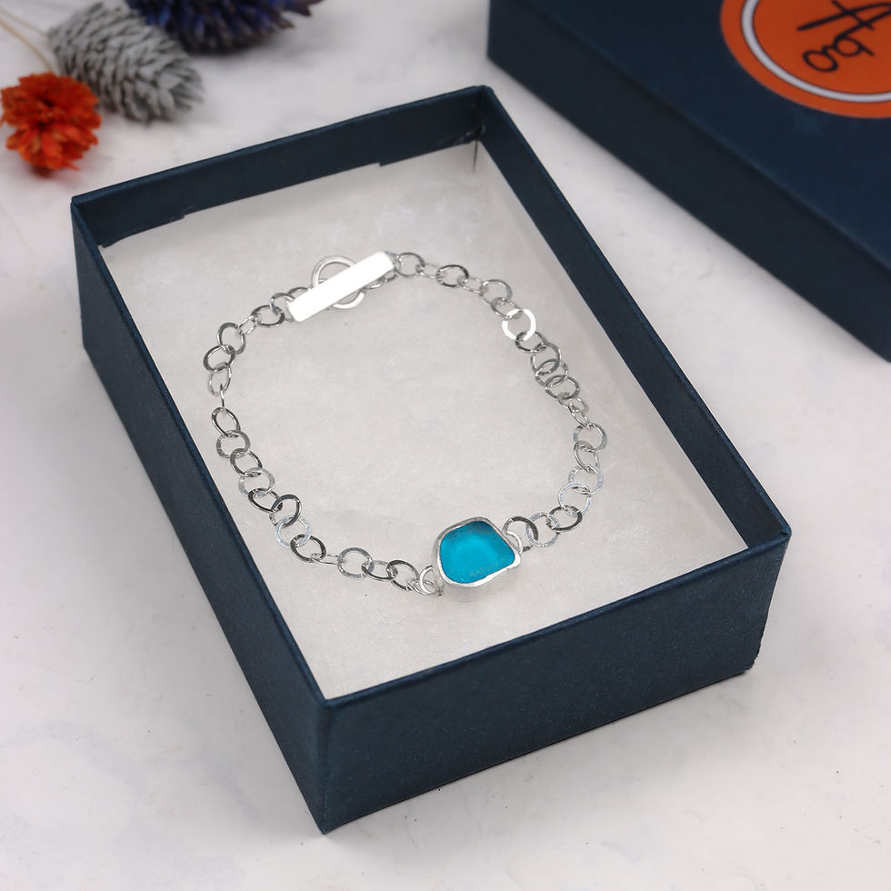 Thumbnail: Aqua blue sea glass and silver chain bracelet in an open dark blue jewellery box on a white background with lid and flowers