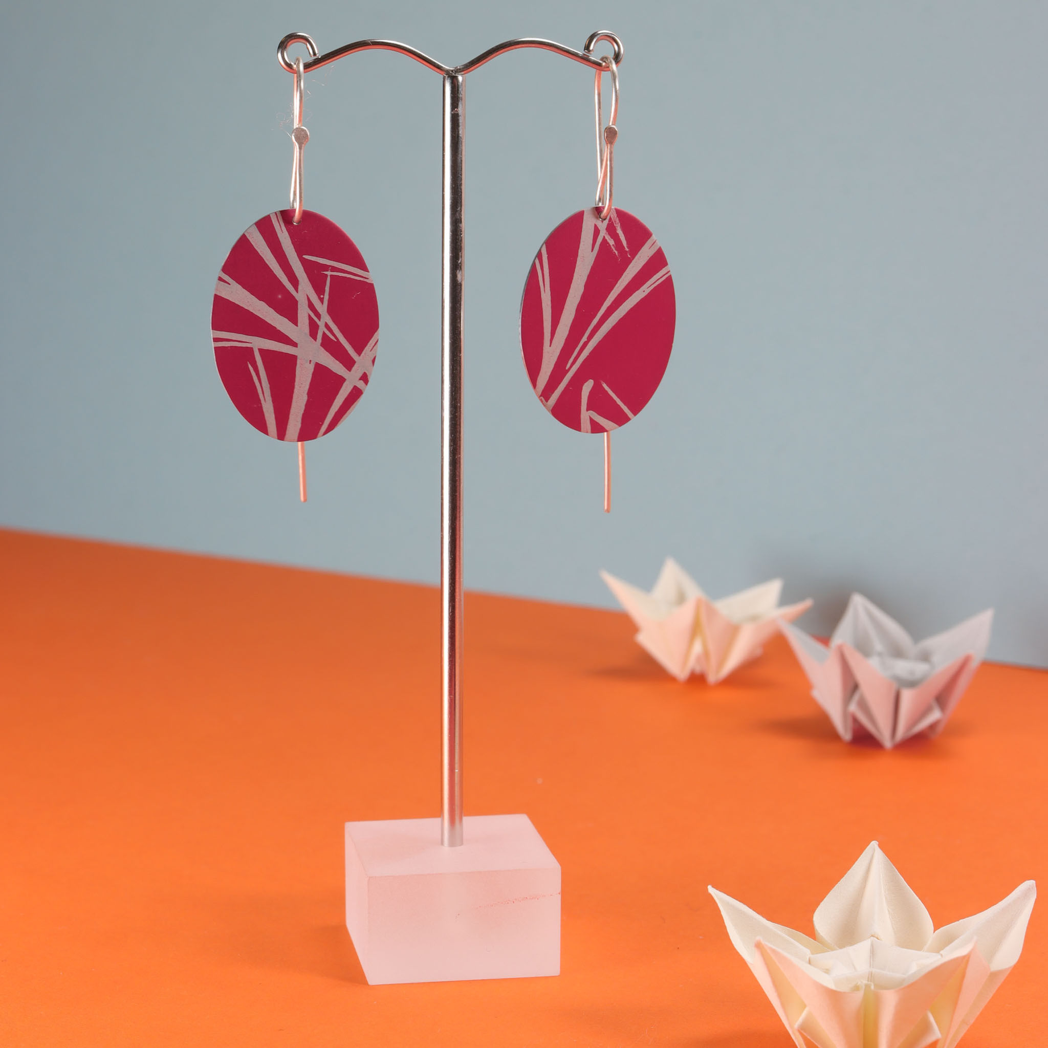 Large magenta pink red and silver oval earrings on an earring stand on an orange background
