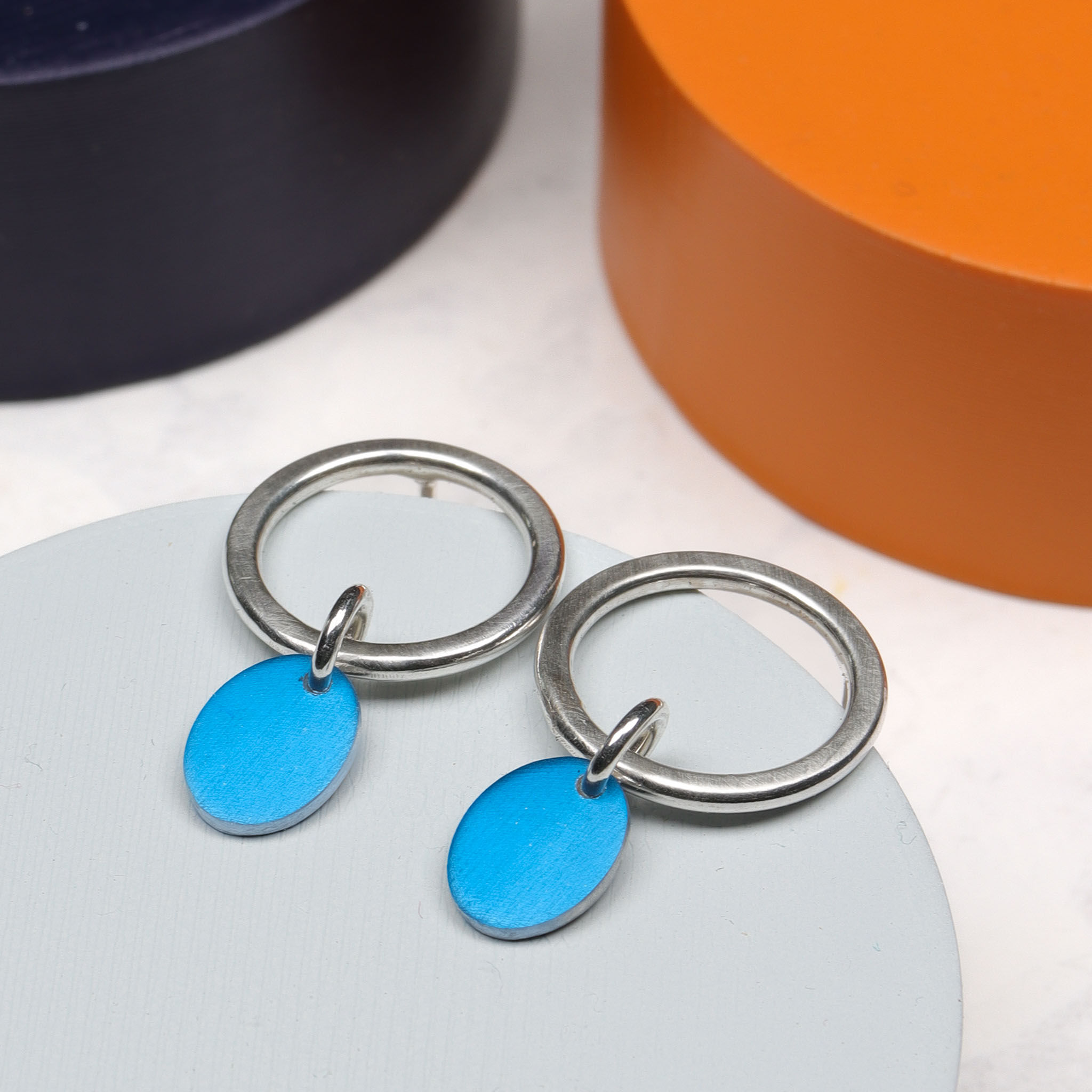 Bright blue oval and silver circle stud drop earrings on a grey plinth with orange and blue plinth behind
