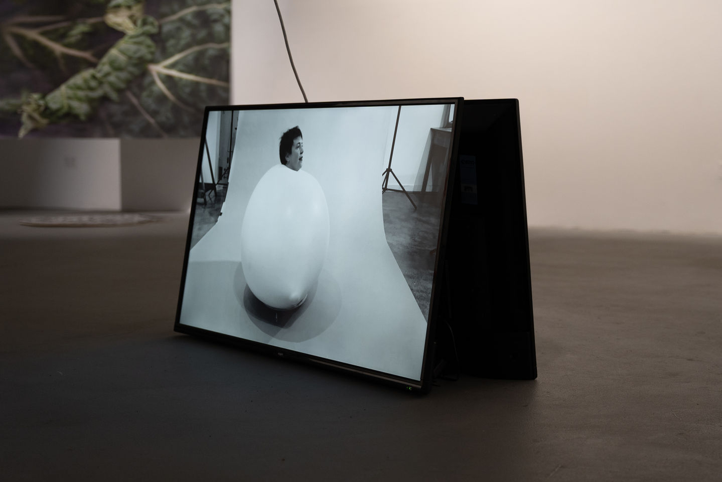 Black-and-white video still showing a large balloon on a studio backdrop with the artist’s head emerging through its surface, hair tousled, breathing fresh air.