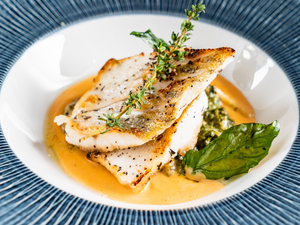 Grilled fish with sprigs of thyme on creamy sauce, set on cooked greens. Served on a blue-rimmed plate, evoking a gourmet presentation.
