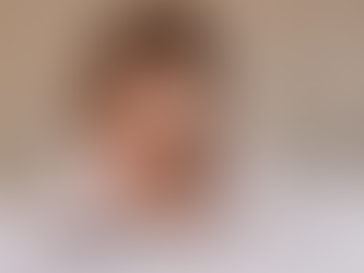 Child with long hair looks surprised while reading a book on a bed. Neutral background, cozy and curious atmosphere, wearing a light top.