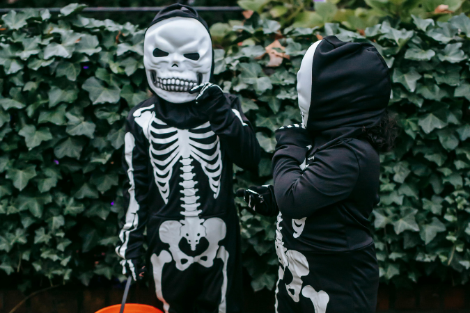 Two children in skeleton costumes stand in front of leafy bushes, holding orange buckets, appearing festive and ready for Halloween.