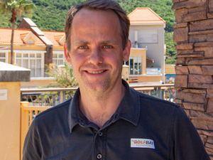 Man smiling in a navy shirt with "GOLF BAY" logo, standing against a stone wall. Background features orange-roofed buildings and greenery.