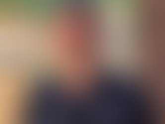 Man smiling in a navy shirt with "GOLF BAY" logo, standing against a stone wall. Background features orange-roofed buildings and greenery.