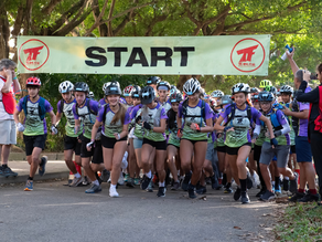 Group of cyclists starting race under a "START" banner in a park. They're wearing helmets and colorful jerseys, with onlookers nearby.