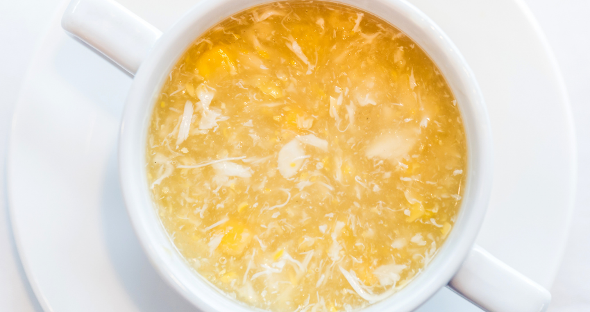 A white bowl of golden chicken and corn soup with visible chicken shreds and corn kernels. The bowl sits on a white saucer.