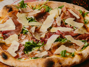 Pizza topped with arugula, prosciutto, and shaved parmesan on a wooden board. Warm, rustic background with red and white cloth detail.
