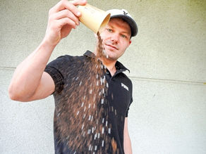 Man in a black shirt and cap pours coffee from a cup indoors against a plain wall. The mood appears casual and focused.