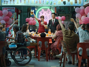 People at a festive dinner raising glasses, celebrating. Pink balloons, a neon sign, and red chairs add lively colors to the scene.