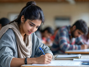 Students in a classroom taking a test. A focused woman writes, surrounded by peers. Warm lighting, casual clothing, and scattered papers.