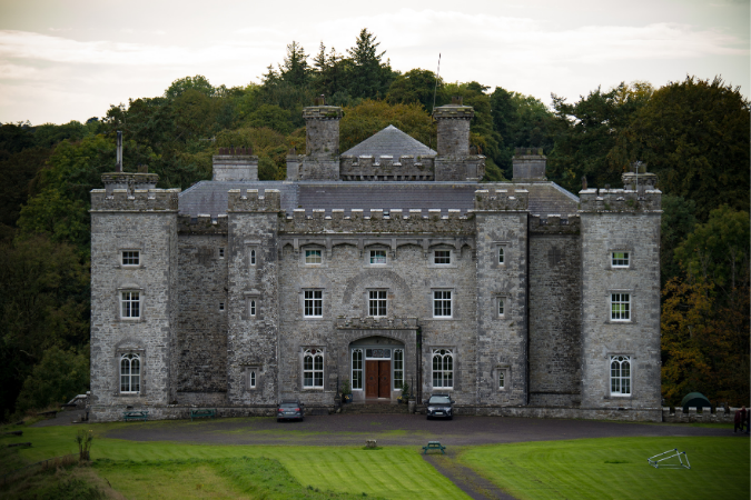 Stone castle with towers set against a backdrop of trees. Two cars parked in front on a grassy lawn. Overcast sky, serene atmosphere.