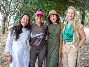 Four women smiling under trees. One wears a pink cap and sports shirt with "Jesus Did It" text. Casual, friendly setting.
