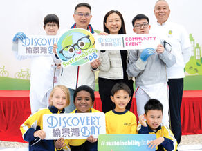A group poses with signs saying "Sustainability in DB" and "Discovery Bay Community Event." Smiling children and adults in vibrant attire.