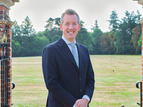 Smiling person in a dark suit stands between brick gate pillars, with a green field and trees in the background under a clear sky.