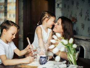 Mom and daughter share a joyful moment, nose-to-nose, while a boy stirs in a jar. Background shows a cozy, patterned room and tulips.