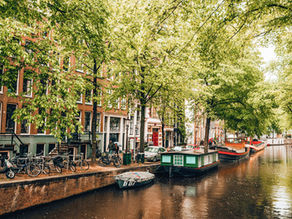 Canal in a city lined with brick buildings, surrounded by lush green trees. Boats and bicycles along the canal, creating a serene atmosphere.