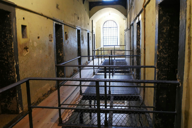 Dimly lit prison corridor with aged yellow walls, metal railings, and barred windows, evoking an eerie and somber atmosphere.