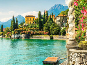 Scenic view of colorful villas surrounded by lush greenery by a lake, with mountains in the background and vibrant flowers in the foreground.