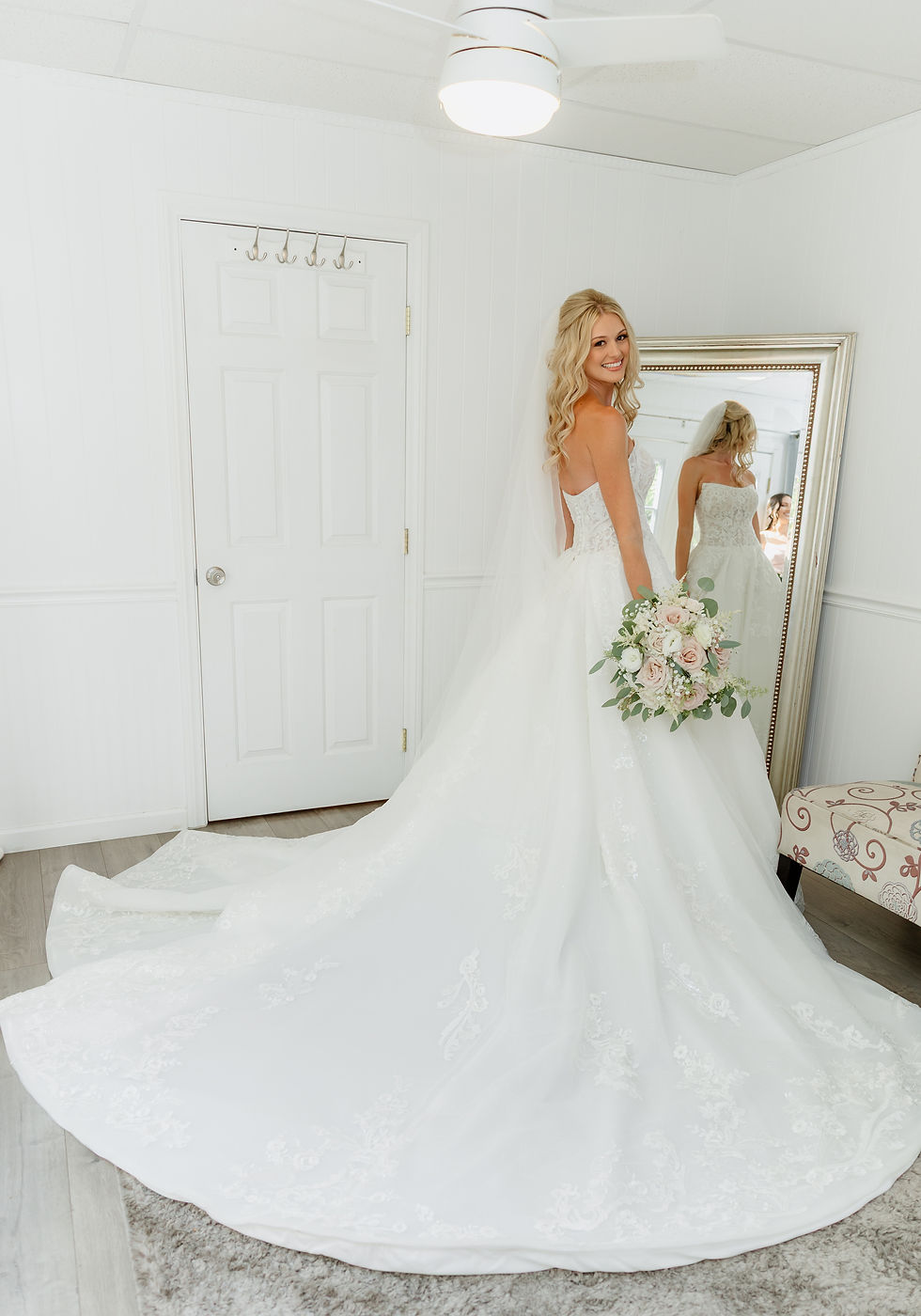 Ashlyn  in her wedding gown holds a floral bouquet, smiling in front of a mirror. White room with a patterned chair; joyful, serene mood.