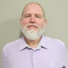 Chris Abbey, CSW, A man with a blonde hair and a white beard wears a purple button down shirt and smiles slightly in front of a beige wall.