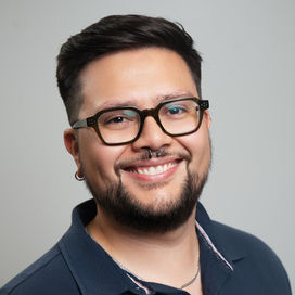 Christopher Bonilla, LCSW, Smiling person with shoulder-length dark hair, glasses, and facial hair wearing a light-colored jacket against a gray background.
