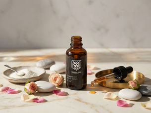 Amber serum bottle labeled Yeshua Skin on marble with spoon, powder, pink roses, petals, and stones. Dropper on golden tray. Calm setting.