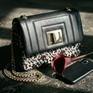 A black leather handbag with a patterned flap and gold chain sits on a textured surface. Nearby is a black phone and a red calla lily.