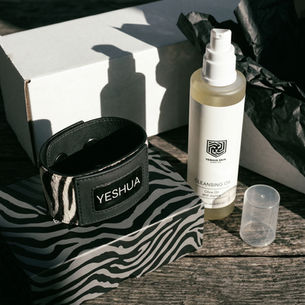 Zebra-patterned box with a black wristband labeled YESHUA, cleansing oil bottle, cap, and white box on a wooden table with sunlight.