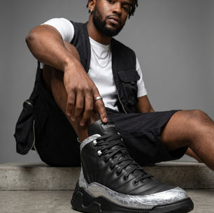 Portrait of a man seated on a concrete floor, pointing at a black high-top sneaker labeled YESHUA against a gray backdrop.