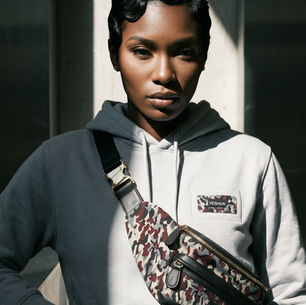 Woman in a white hoodie with a camo crossbody bag labeled YESHUA, lit by a bright window, looking calmly at the camera.