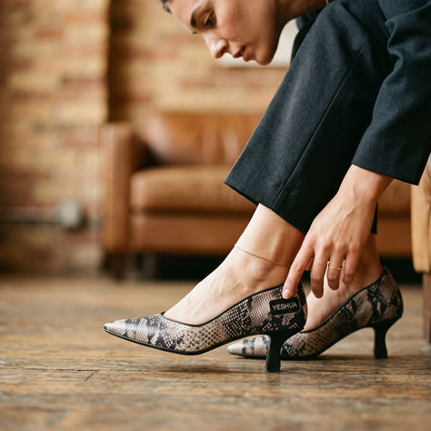 Woman in black pants adjusts snakeskin heels labeled YESHUA in a warm brick-and-leather room.