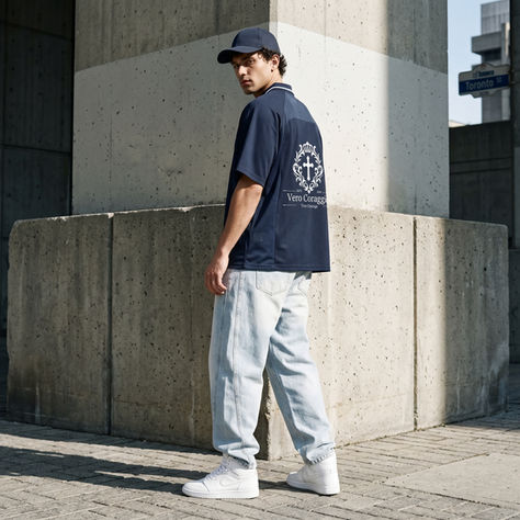 Man in a navy shirt with "Vero Coraggio" on the back, light jeans, white sneakers, and a cap stands by a concrete wall on Toronto St.