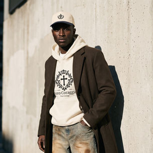 Young man in cap, hoodie and coat leans by a sunlit concrete wall, wearing a Vero Coraggio True Courage sweatshirt.