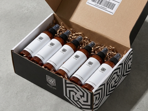 Open box with six brown bottles on shredded paper. Box has black and white geometric patterns and shipping label on a gray surface.