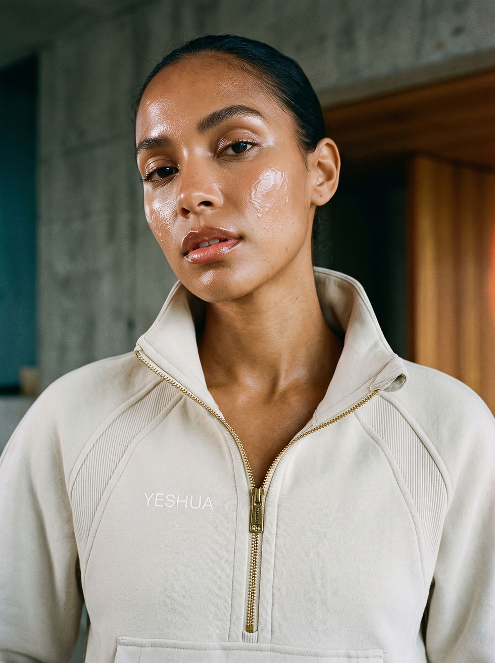 Close-up of a woman in a cream zip-up jacket with YESHUA text, looking calmly at the camera indoors.