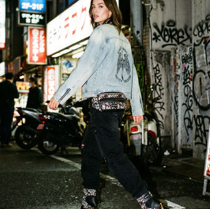 Woman in denim jacket and heels walking on graffiti-lined street at night, surrounded by neon signs and motorcycles.