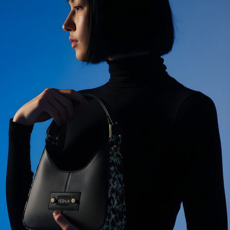 A person in a black turtleneck holds a stylish black bag labeled "YESHUA" against a blue background, creating a sleek, moody look.