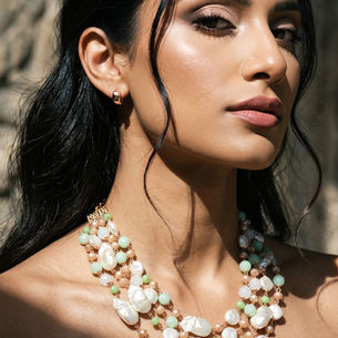 Close-up of a woman with dark hair and gold earrings, wearing a pastel beaded necklace, lit by warm sunlight against a blurred stone background.