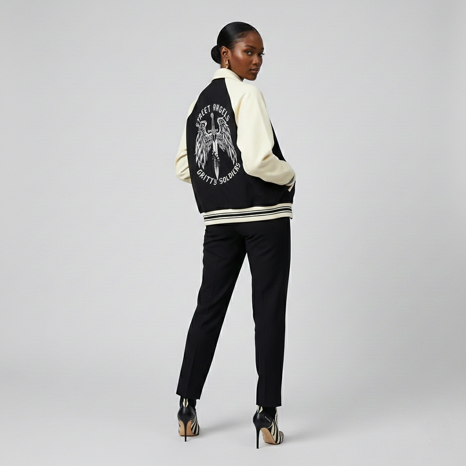 A person wearing a black and cream varsity jacket with an eagle and sword graphic on the back, black pants, and black and white patterned heels against a light gray background.