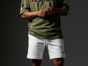 Man in green "YESHUA" tee and white shorts stands confidently. Wears black hat and sunglasses. Gray background, casual style.