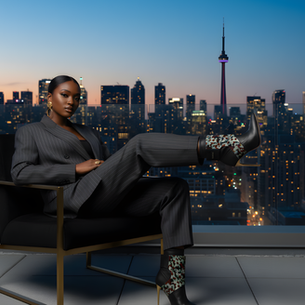Woman in a pinstripe suit reclines in a chair on a balcony, overlooking a city skyline at dusk. Stylish boots and bold earrings accent her look.
